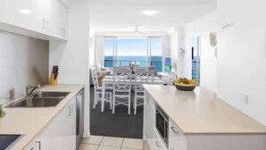 A modern kitchen with a central island, a dining area with white chairs, and a view of the sea through large windows. - Chevron Renaissance Ocean View (Surfers Paradise)