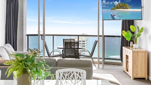 Un balcón con vistas al océano, un comedor con mesa y sillas, y una pantalla de televisión mostrando un paisaje tropical.