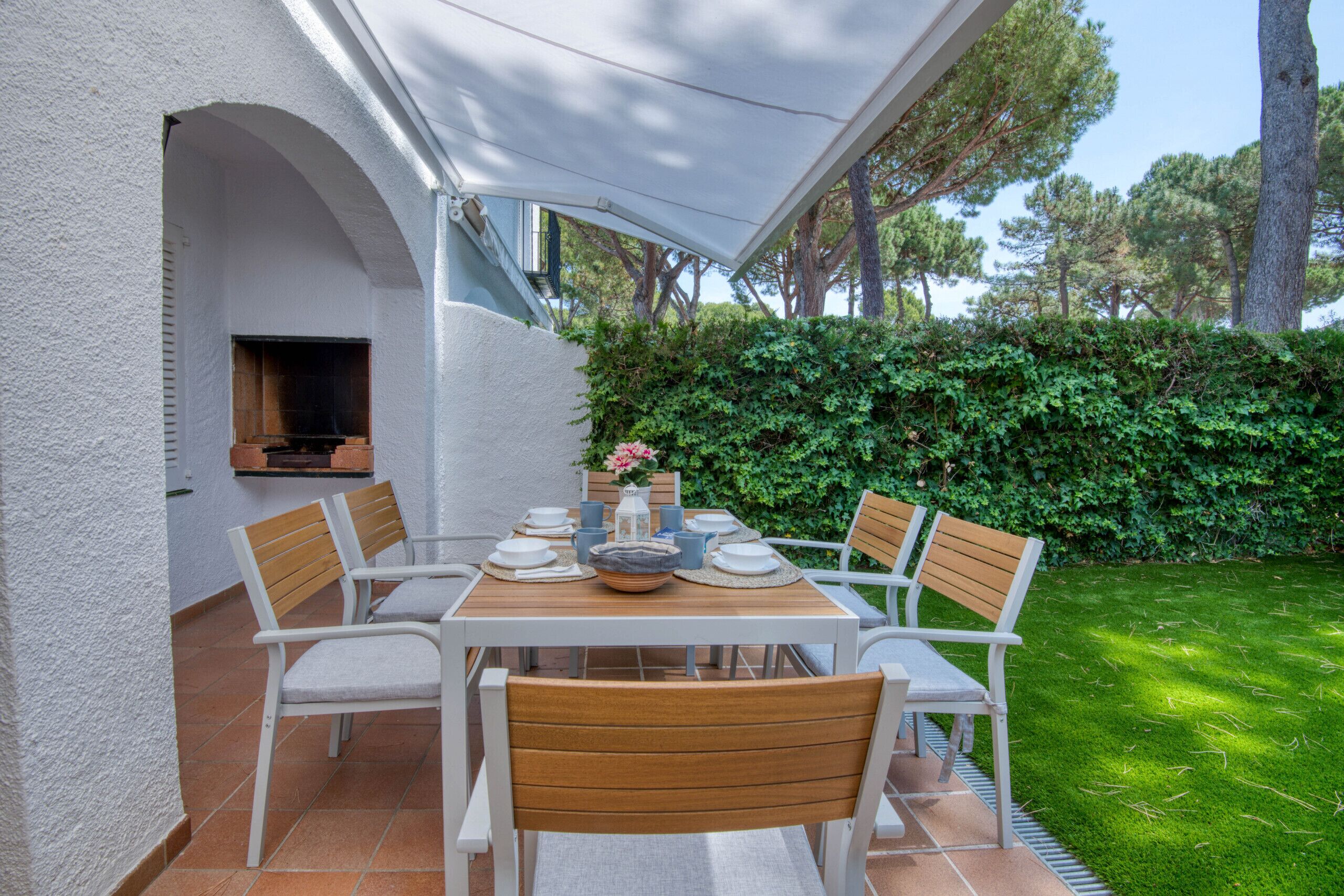 A patio with a table set for four, surrounded by white chairs, a TV mounted on the wall, and a hedge in the background.