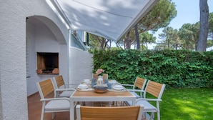Eine Terrasse mit Esstisch für vier Personen, umgeben von weißen Stühlen, einem an der Wand befestigten Fernseher und einem Hecken im Hintergrund.