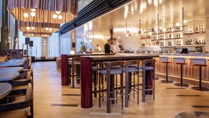 Zona de bar moderna con mesas y taburetes de madera, una barra con un cantinero y estantes llenos de botellas.