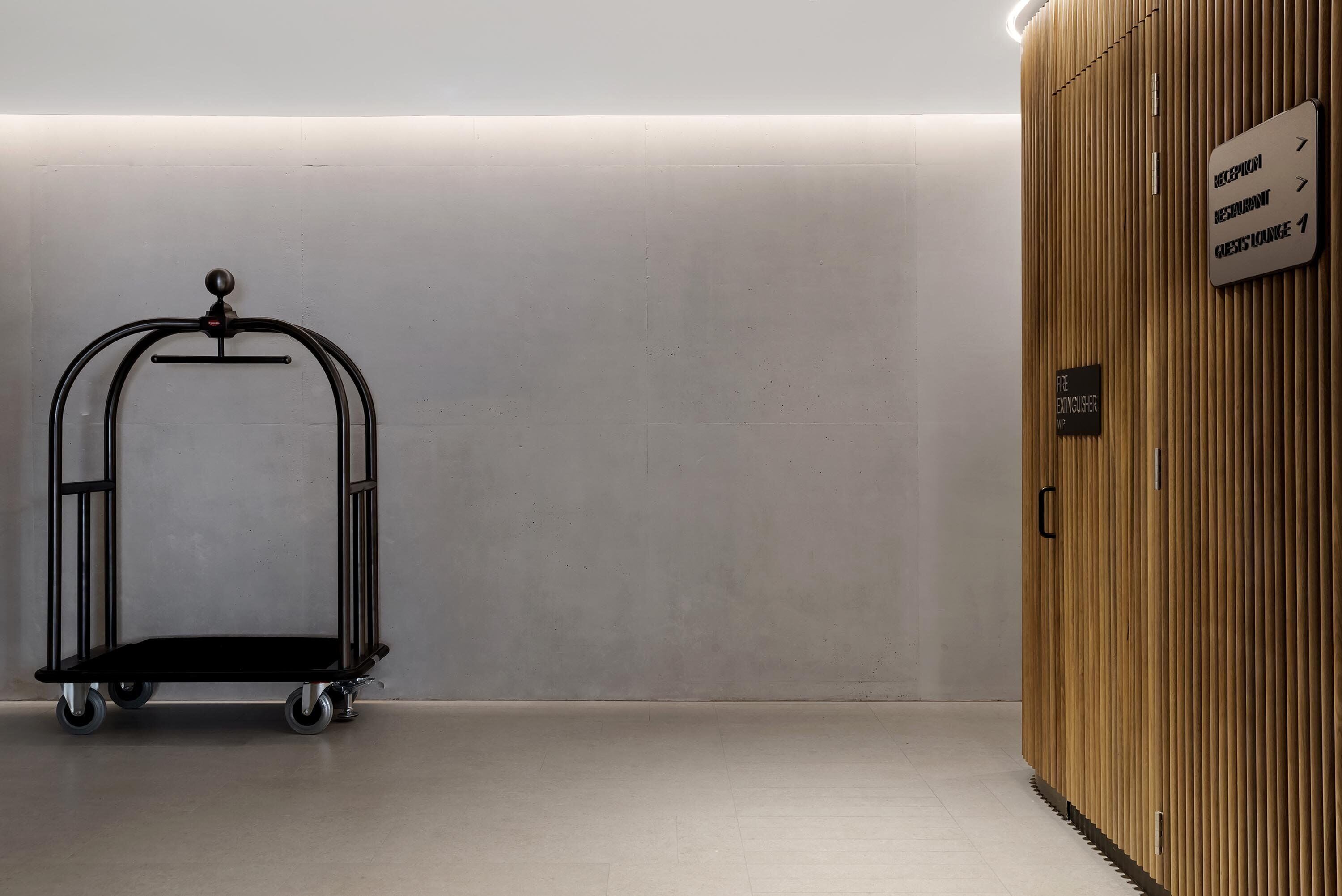 A clean, modern hotel lobby with a black luggage cart, a wooden reception desk, and directional signs for the restaurant and guest lounge.