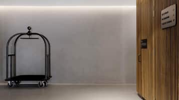 A clean, modern hotel lobby with a black luggage cart, a wooden reception desk, and directional signs for the restaurant and guest lounge.
