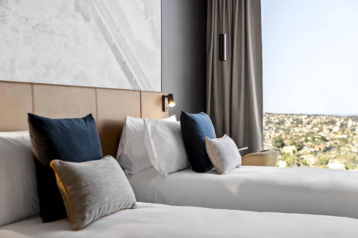 A modern hotel room with a large bed, multiple pillows, a headboard with a light fixture, and a view of a cityscape through large windows.