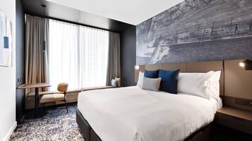 Habitación de hotel moderna con una cama grande, un escritorio y una silla. Hay un ventanal con cortinas, una obra de arte en la pared y un cabecero con almohadas.