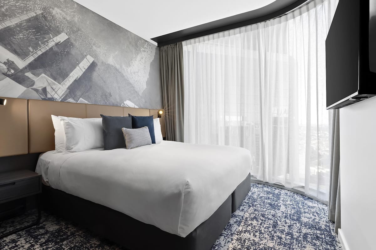 A modern hotel room with a large bed, a headboard with a geometric wall art, a flat-screen TV, and a large window with sheer curtains.