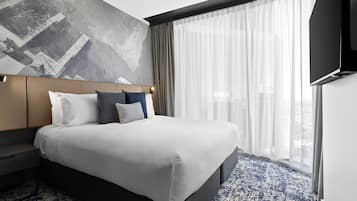 Ein modernes Hotelzimmer mit einem großen Bett, einem Kopfteil mit geometrischer Wandkunst, einem Flachbildfernseher und einem großen Fenster mit transparenten Vorhängen.