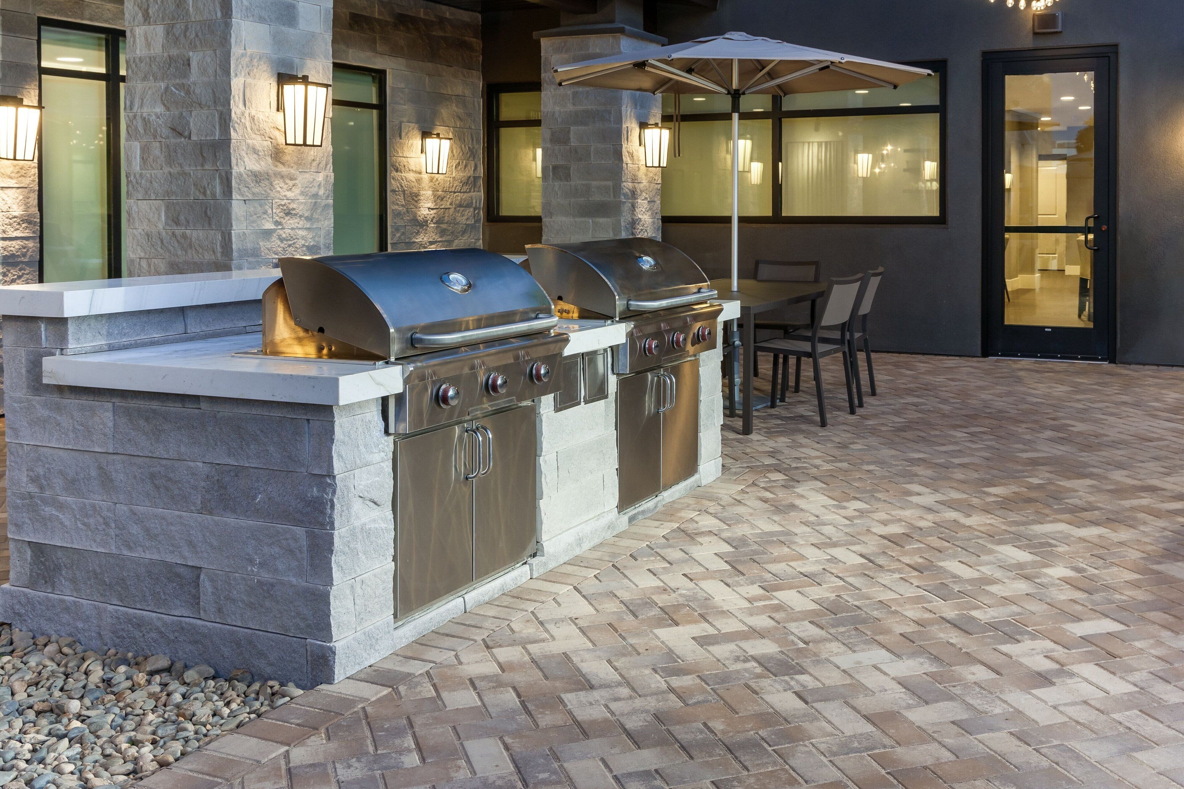 An outdoor kitchen with a stone countertop and stainless steel appliances, including a grill and a sink. There are two wall-mounted lights and a table with chairs.