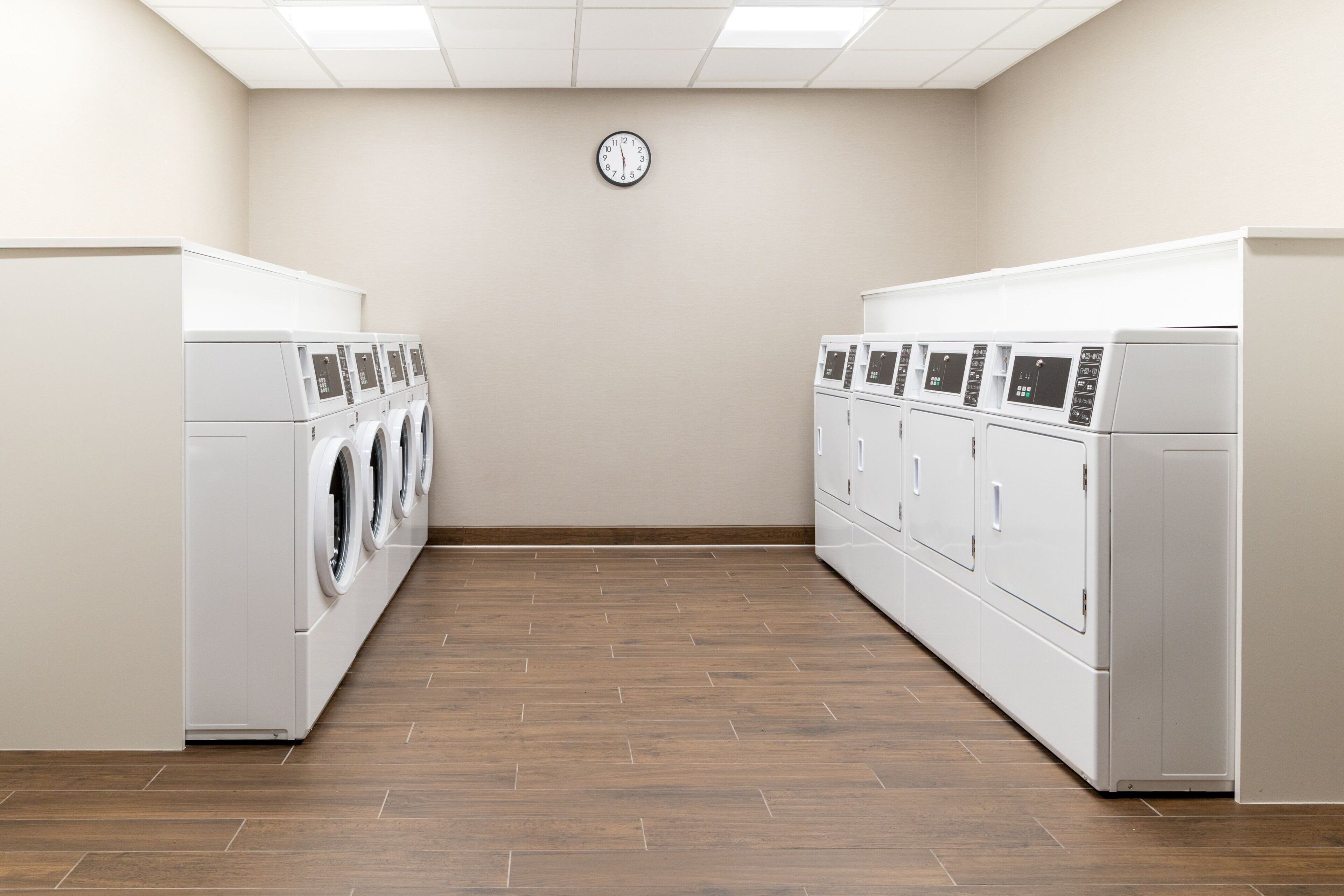 A laundry room with rows of washing machines and dryers, a clock on the wall, and wooden flooring.