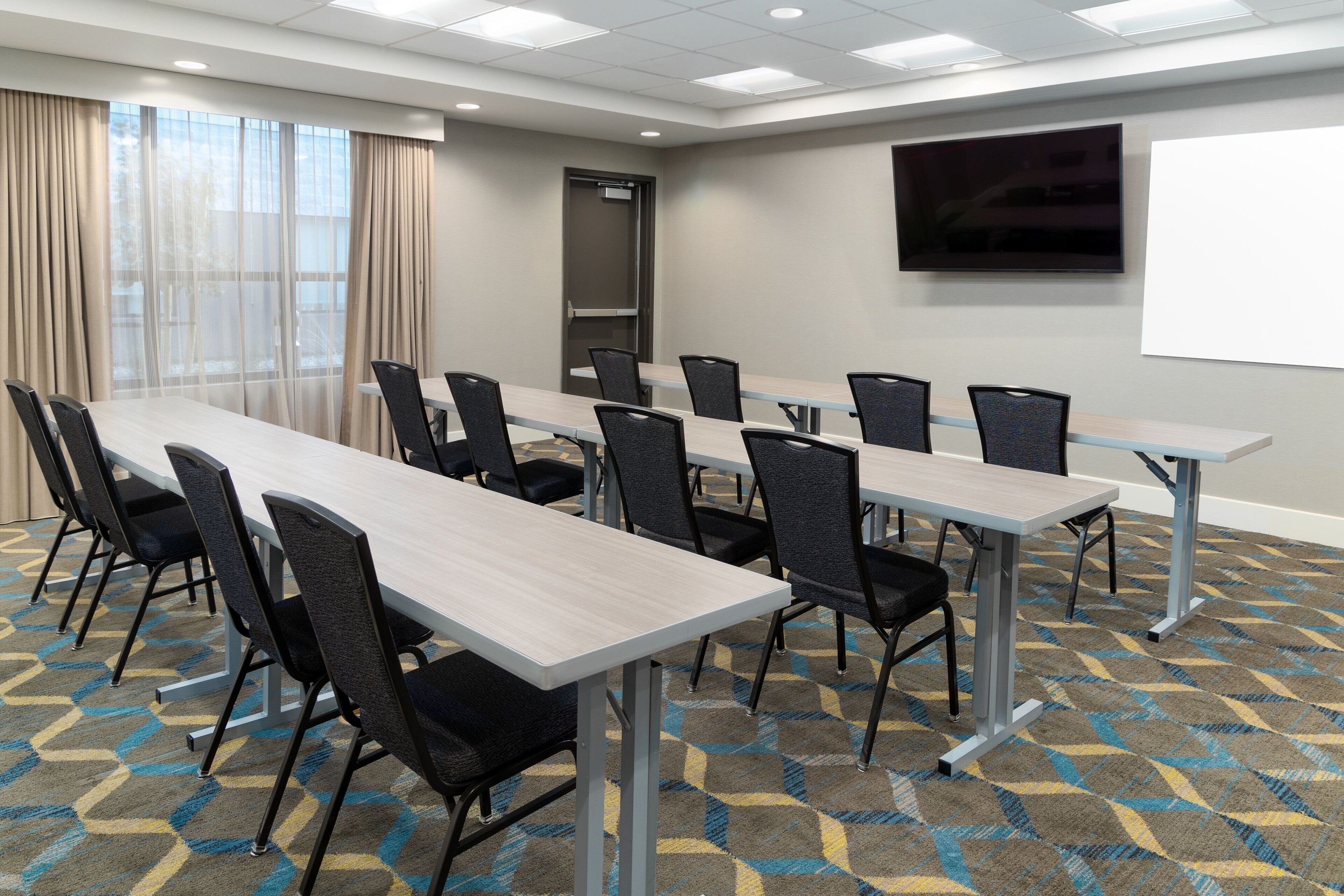 A conference room with a long table, multiple chairs, a projector screen, and a large window with curtains.
