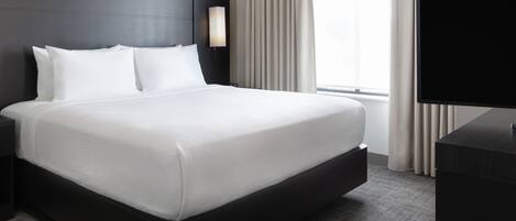 Habitación de hotel con una cama grande, una mesita de noche, un televisor de pantalla plana y un ventanal con cortinas.