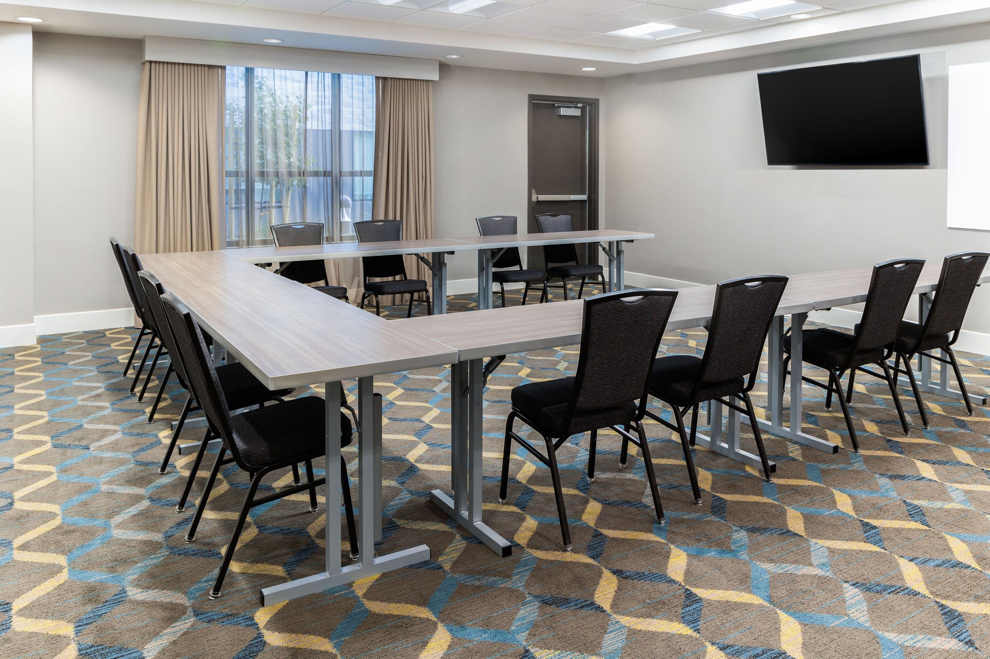 A conference room with a long table, black chairs, a patterned carpet, a large window with curtains, and a flat-screen TV on the wall.