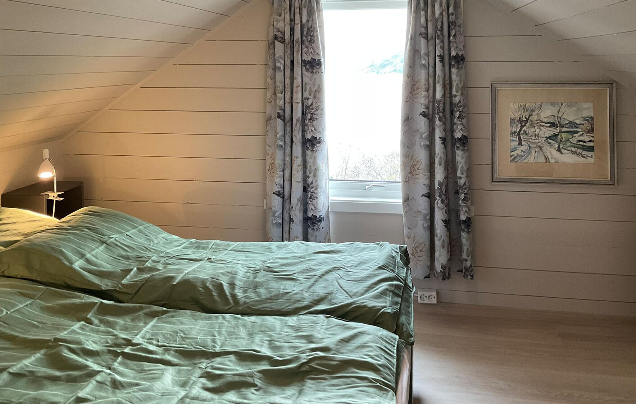 Une chambre avec un plafond en pente, un lit avec une literie verte, une lampe de chevet, des rideaux à motifs floraux et un tableau encadré accroché au mur.