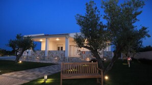 A modern house with a well-lit porch, a wooden bench, and a tree in the foreground.