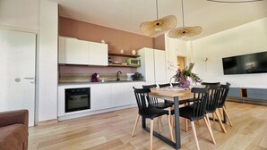 A modern kitchen with white cabinets, a microwave, and a sink. There is a dining table with chairs and a TV mounted on the wall.
