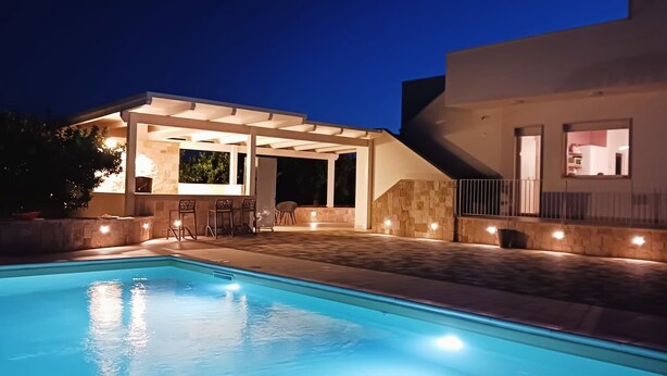 A swimming pool with a covered patio area, stone walls, and a modern building with a balcony.