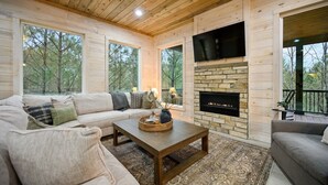 Un salon chaleureux avec une cheminée en pierre, un plafond en bois et de grandes fenêtres donnant sur la forêt.