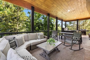 Relax on the patio with a sectional sofa, coffee table, and rocking chairs, surrounded by lush greenery and a wooden deck.