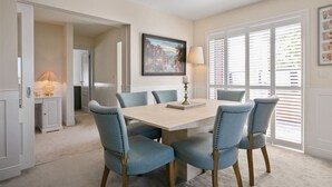A dining room with a wooden table and six blue upholstered chairs. There is a painting on the wall and a window with blinds. - Luxurious 2-Bed Apartment - Jun-Aug Weekly Rate (Rathfarnam Dublin 14)