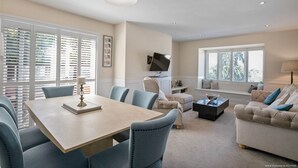 A modern living room with a dining area, large windows with blinds, a flat-screen TV mounted on the wall, and a comfortable seating arrangement. - Luxurious 2-Bed Apartment - Jun-Aug Weekly Rate (Rathfarnam Dublin 14)