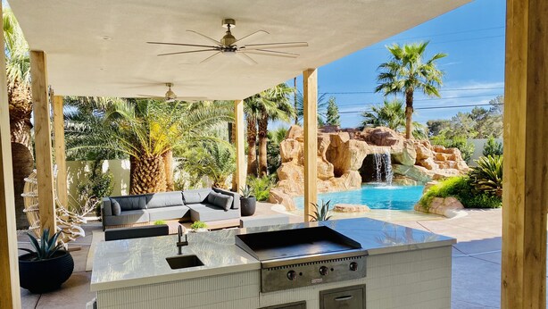 A covered outdoor kitchen area with a built-in island, seating, and a view of a natural landscape with a pool and waterfall. - Resort Living Las Vegas (Las Vegas)