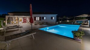 Une piscine avec des chaises longues et des parasols, un patio couvert et une maison en arrière-plan.