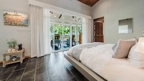 Quarto com cama grande, porta de correr de vidro para uma varanda, ventoinha no teto e um quadro na parede.