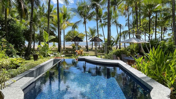 Piscina com bordas azulejadas, rodeada por vegetação exuberante e palmeiras, com vista para a praia ao fundo.