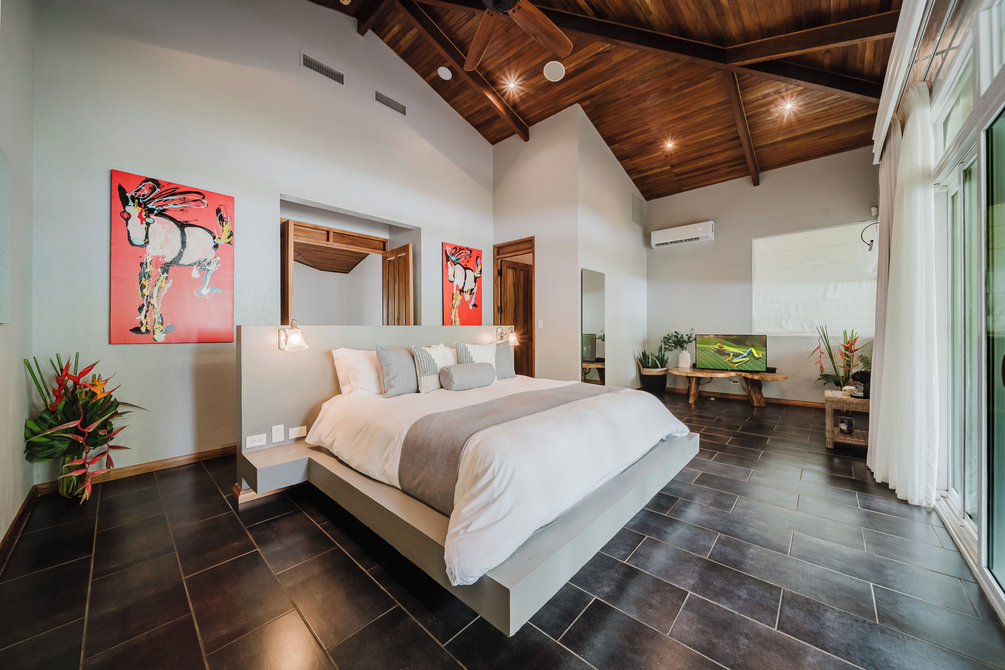 A modern bedroom with a large bed, wooden ceiling beams, and artwork on the walls.