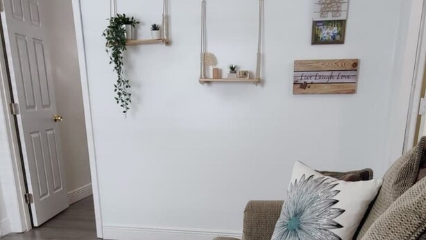 Una habitación con un sofá color beige, un cojín decorativo, un estante de madera con plantas y un cartel enmarcado en la pared.