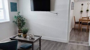 Una sala de estar moderna con un televisor de pantalla plana montado en la pared, un comedor con muebles de madera y una planta en maceta sobre una mesita auxiliar.