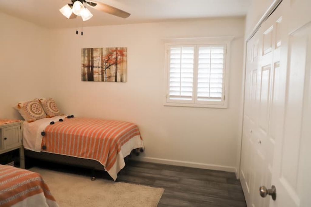 A bedroom with a bed, a nightstand, a ceiling fan, a window with blinds, and a door.