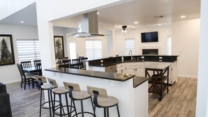 A modern kitchen with a central island, bar stools, a dining area with chairs, and a TV mounted on the wall.