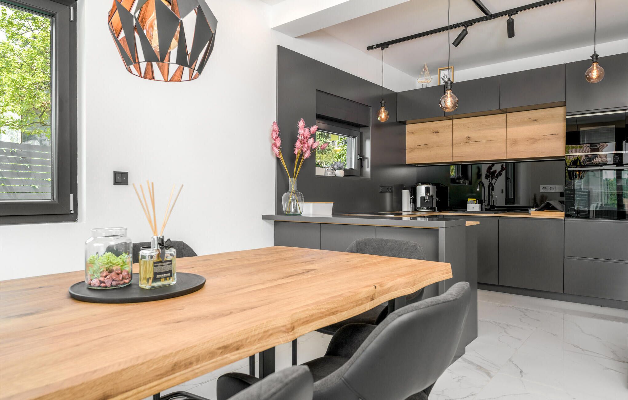 Une cuisine moderne avec des meubles de couleur sombre, une table à manger en bois et des luminaires suspendus.