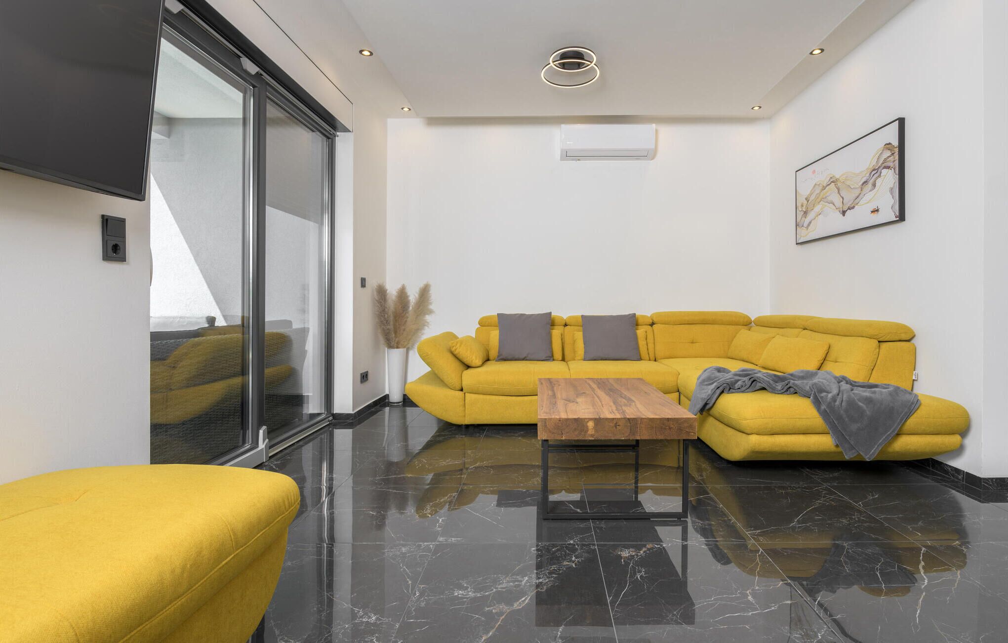 A modern living room with a yellow sofa, a wooden coffee table, and a glass door leading to a balcony.