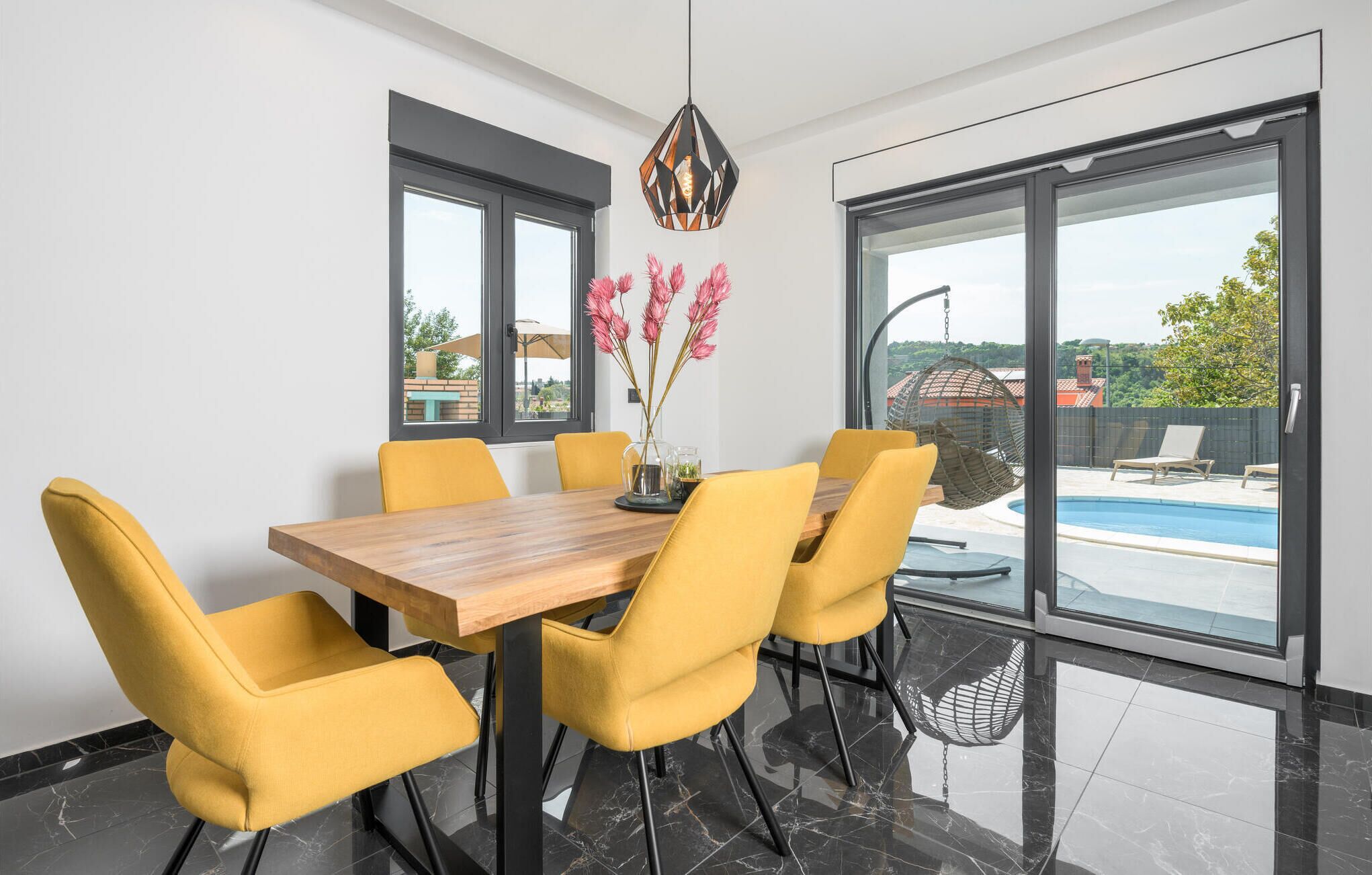 Comedor con una mesa de madera y sillas amarillas, ventanas amplias con vista a una piscina y zona de estar al aire libre.