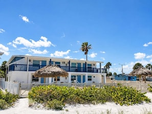 A two-story building with a white and blue exterior, a thatched roof, and a sandy beach in the foreground. - 🌊Chateau 3E - Beach front complex! Pool is Closed🌊 (Belleair Beach)