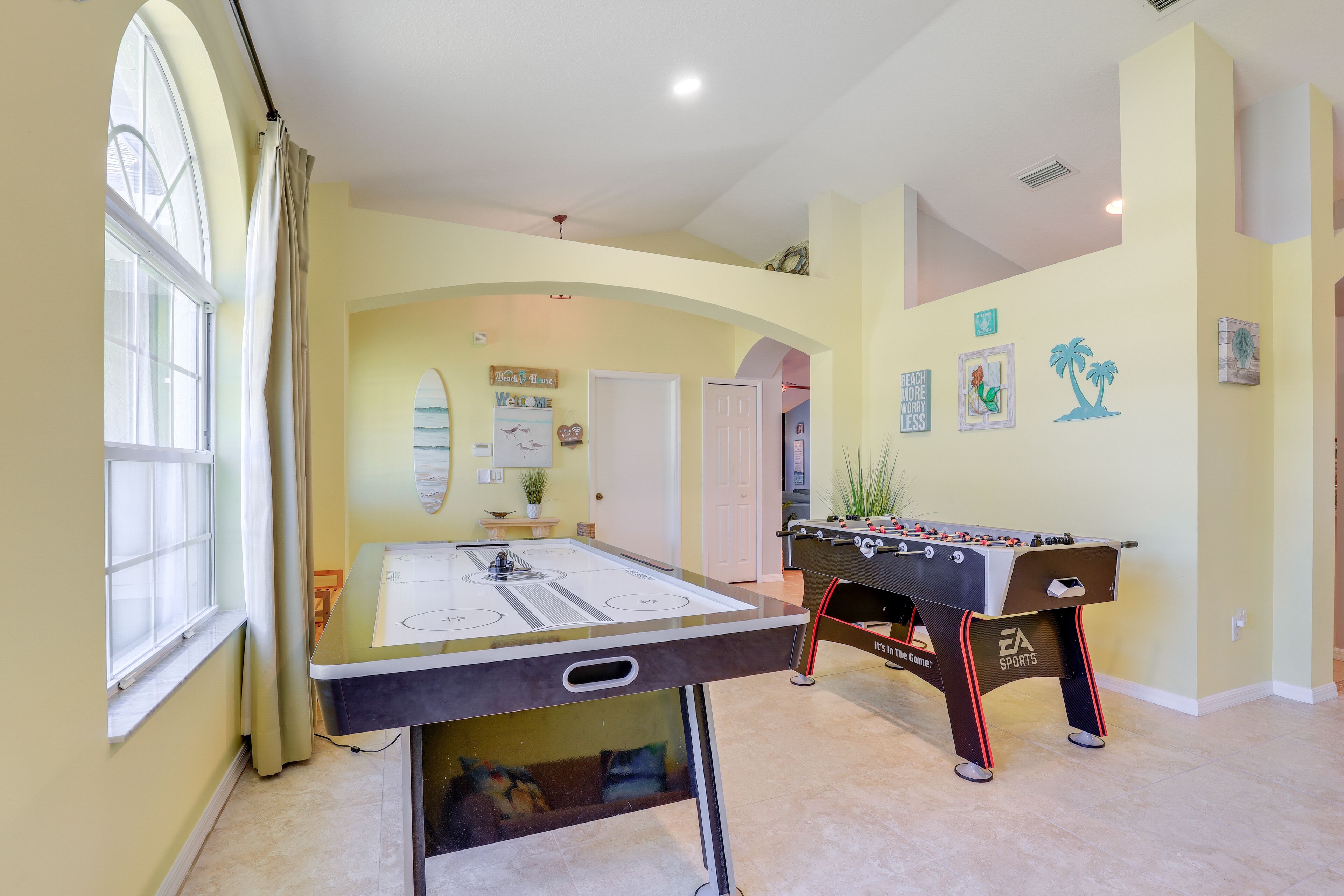A room with a foosball table, a ping pong table, and a large window with curtains.