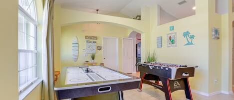 A room with a foosball table, a ping pong table, and a large window with curtains.