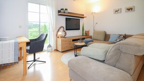 Ett modernt vardagsrum med en soffa, ett litet bord, en tv som är monterad på väggen och ett fönster med utsikt över grönområden.