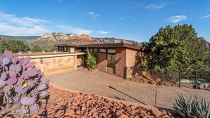 A modern building with a flat roof, large windows, and a brick exterior. It is surrounded by a landscaped area with cacti and a tree. - Walk to Uptown with Breathtaking Views and HotTub! (Sedona)