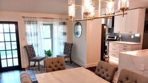Ein Essbereich mit einem Marmortisch und gestopften Stühlen, eine Küche mit weißen Schränken und ein großes Fenster mit durchscheinenden Vorhängen.