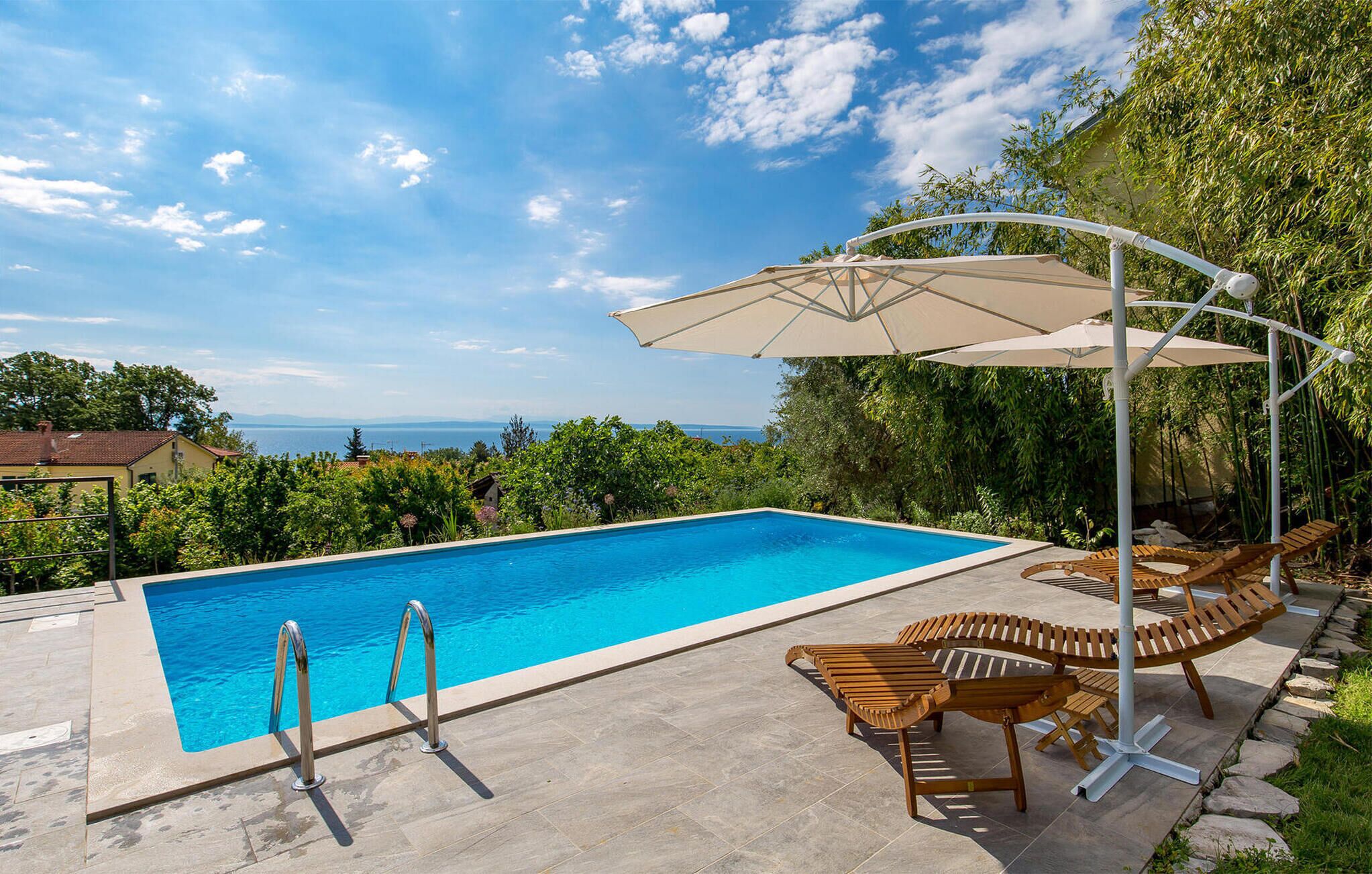 Une piscine rectangulaire à l’eau d’un bleu limpide, entourée d’une terrasse en pierre. On y trouve des chaises longues en bois et un parasol blanc doté d’une toile.