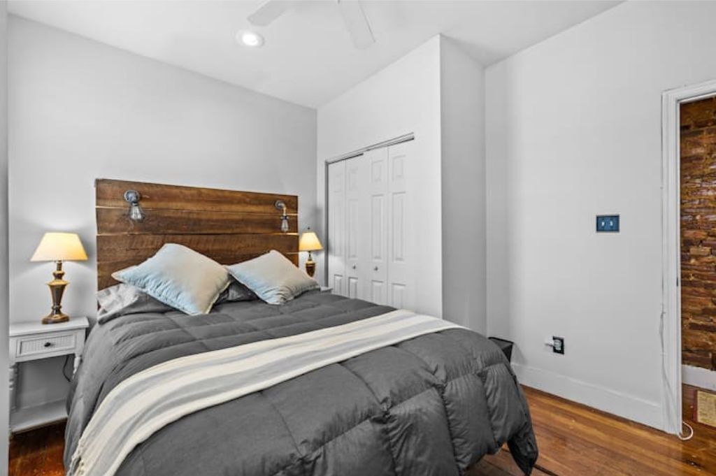 A bedroom with a large bed, a wooden headboard, two bedside lamps, a white door, and a brick wall.