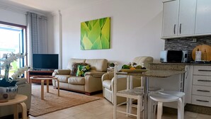 Una sala de estar moderna con un sofá, una mesa de centro y un televisor. Hay una zona de cocina con microondas y un frutero.