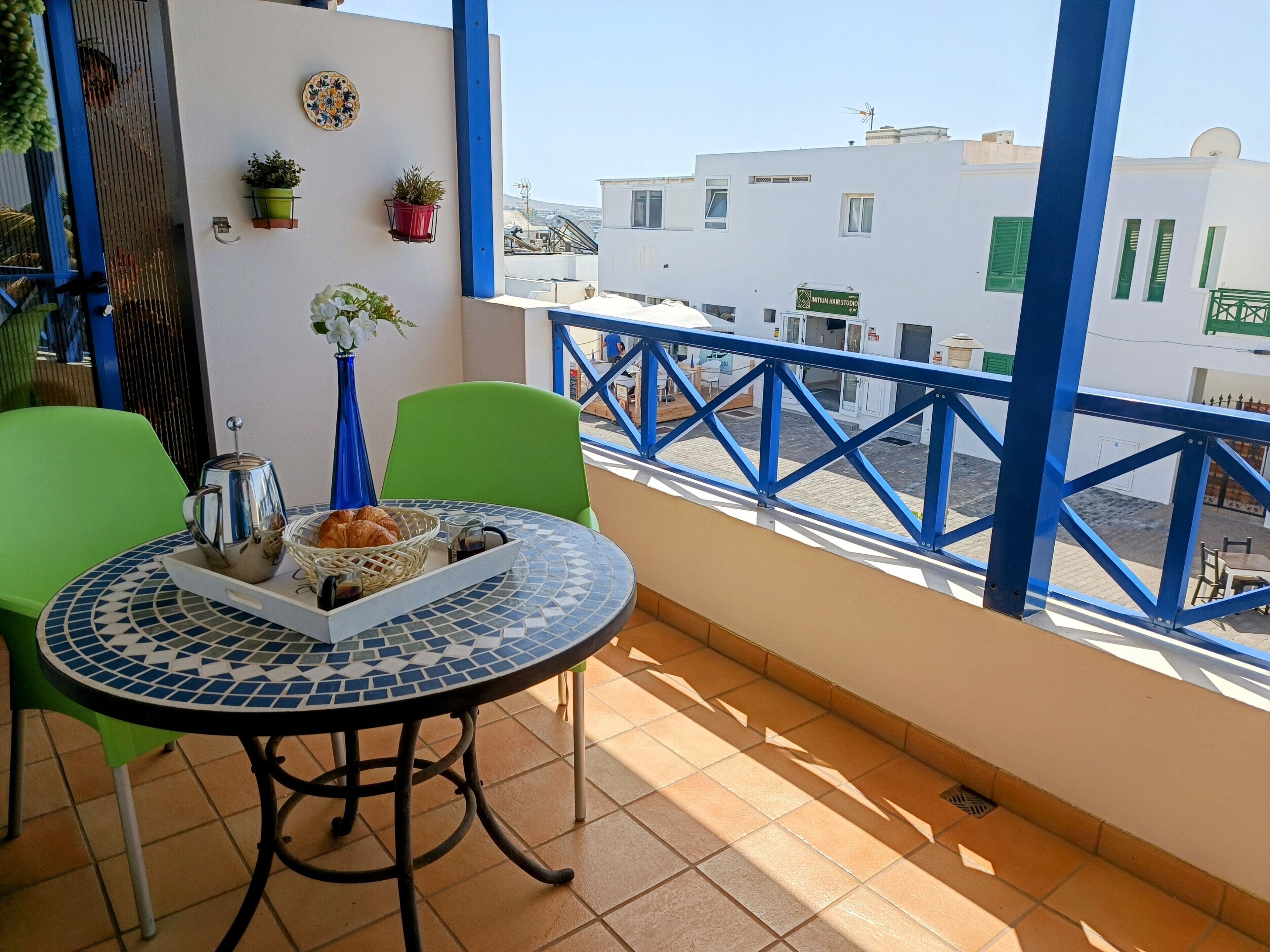 Un balcon avec un sol carrelé, une table ronde entourée de chaises, une théière et un panier de croissants.
