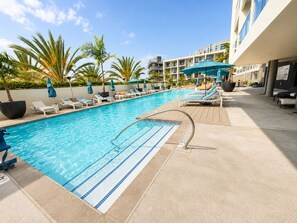 A swimming pool with lounge chairs and umbrellas, surrounded by palm trees and a building with balconies.