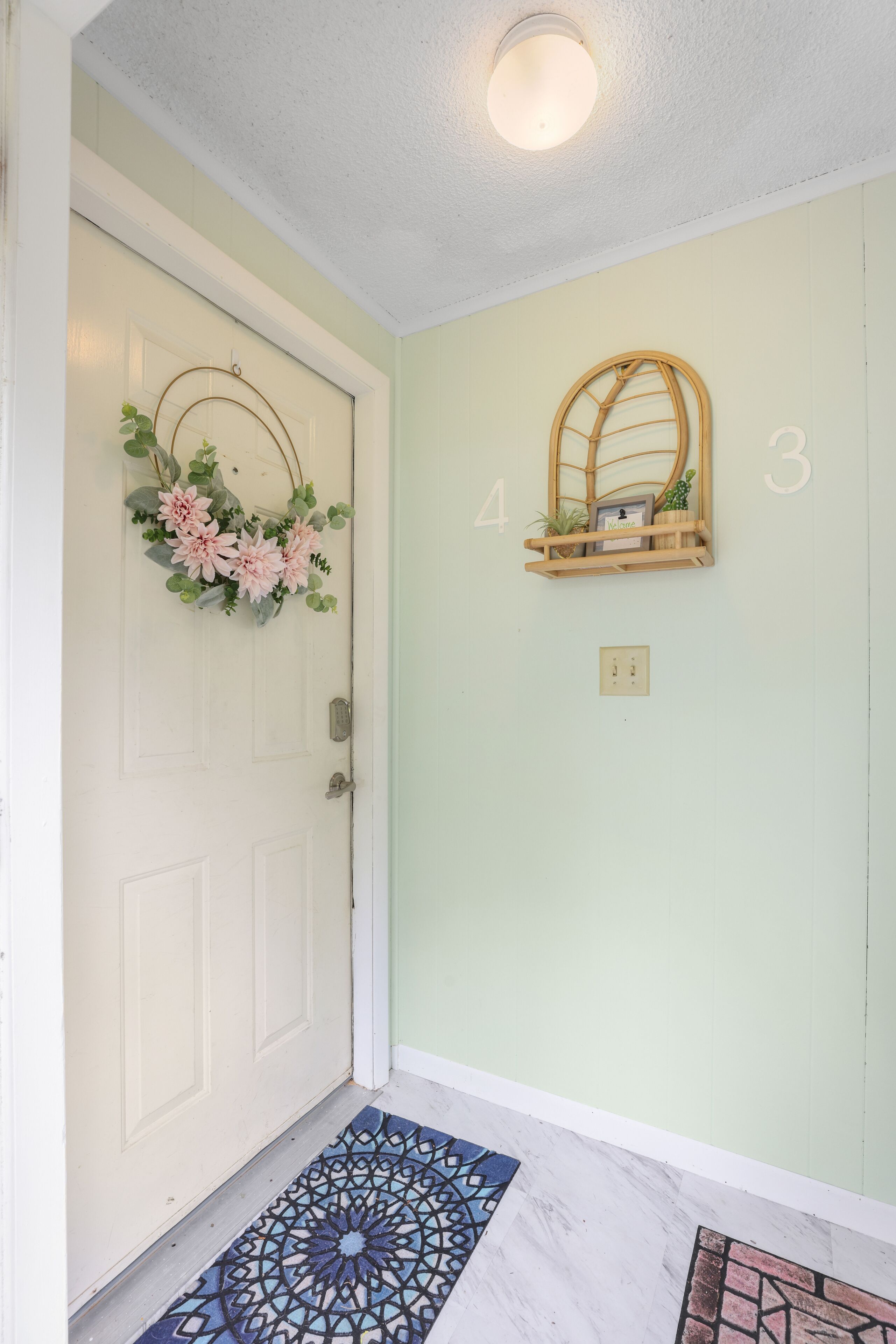 A door with a floral wreath, a small shelf with a mirror and a picture frame, and a patterned doormat.