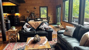 L’intérieur chaleureux d’un chalet avec une cheminée, des canapés en cuir, une table basse en bois et de grandes fenêtres offrant une vue sur l’extérieur.