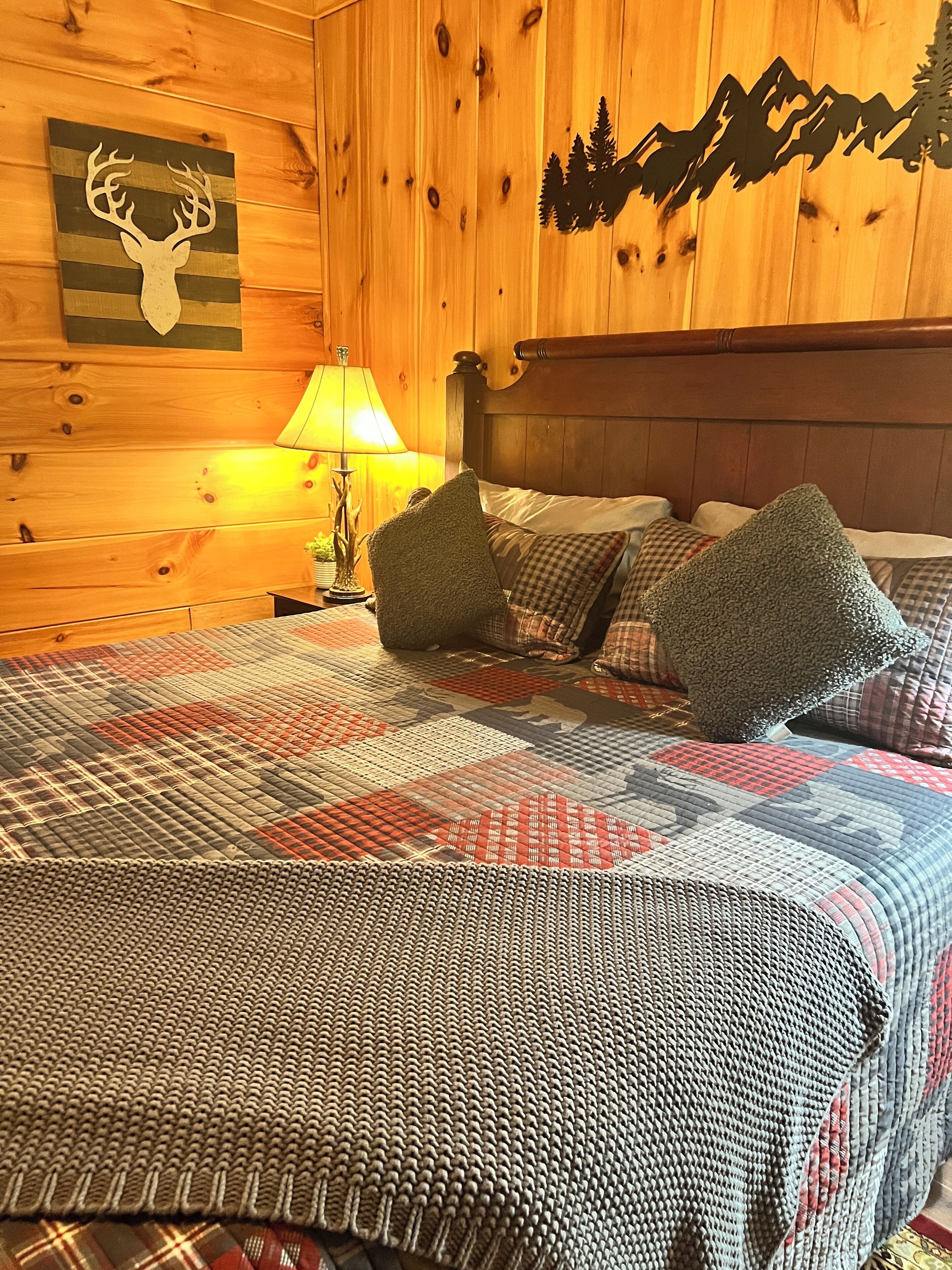 A wooden cabin room with a bed, pillows, a lamp, and a wall decoration of a deer head and mountains.